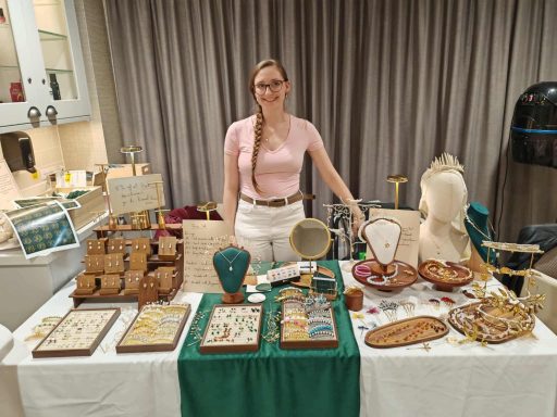 A woman stands behind a table displaying various handmade jewellery and crafts.